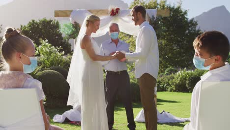 caucasian bride groom and wedding officiant wearing face mask standing at outdoor altar