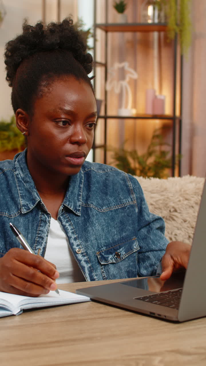 African american young woman studies online with laptop and notebook takes write notes at home table
