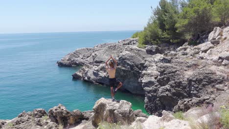 una persona haciendo la pose de yoga del árbol junto a una pequeña cala rocosa en portugal