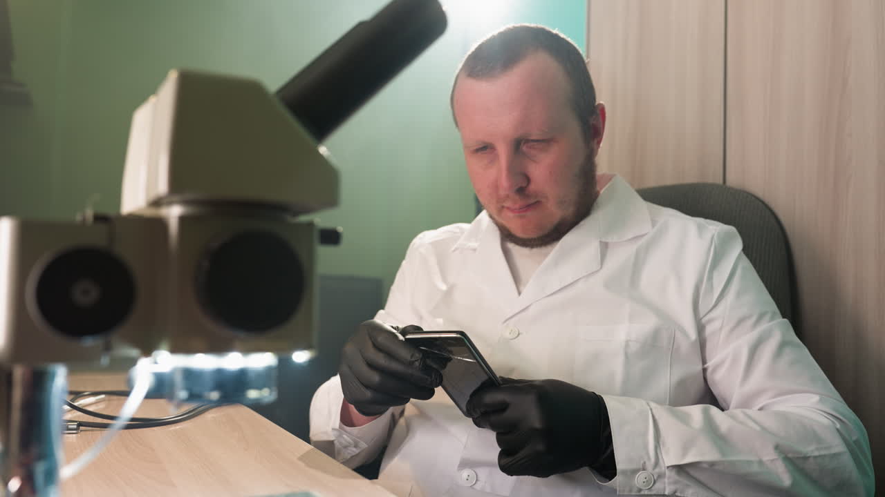 una vista en primer plano de un técnico en una bata de laboratorio blanca y guantes negros examinando cuidadosamente un teléfono inteligente con una pantalla de grieta mientras está sentado en un escritorio en un laboratorio
