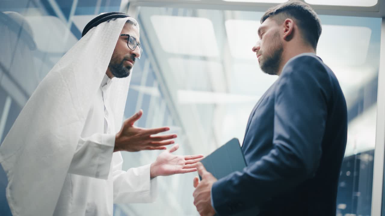 un hombre de negocios hablando con un socio de inversión árabe mientras viaja en un ascensor de vidrio a su oficina en un moderno centro de negocios.