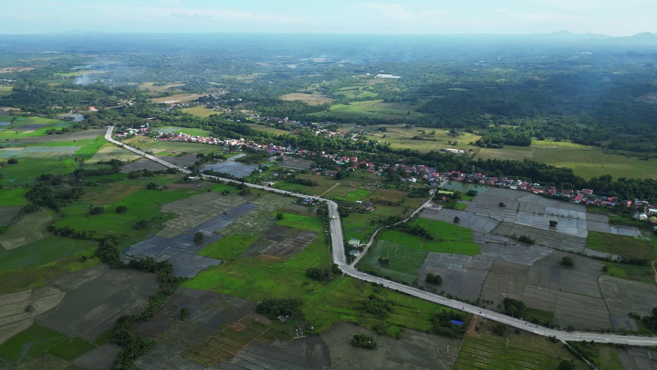 ภาพมุมสูงแบบเคลื่อนไหวช้าของถนนในชนบทข้างทุ่งนาสีเขียวและบ้านเรือนที่กระจัดกระจายในเวลากลางวัน ณ เมืองมูซอน ไนก์ คาไวต์ ฟิลิปปินส์