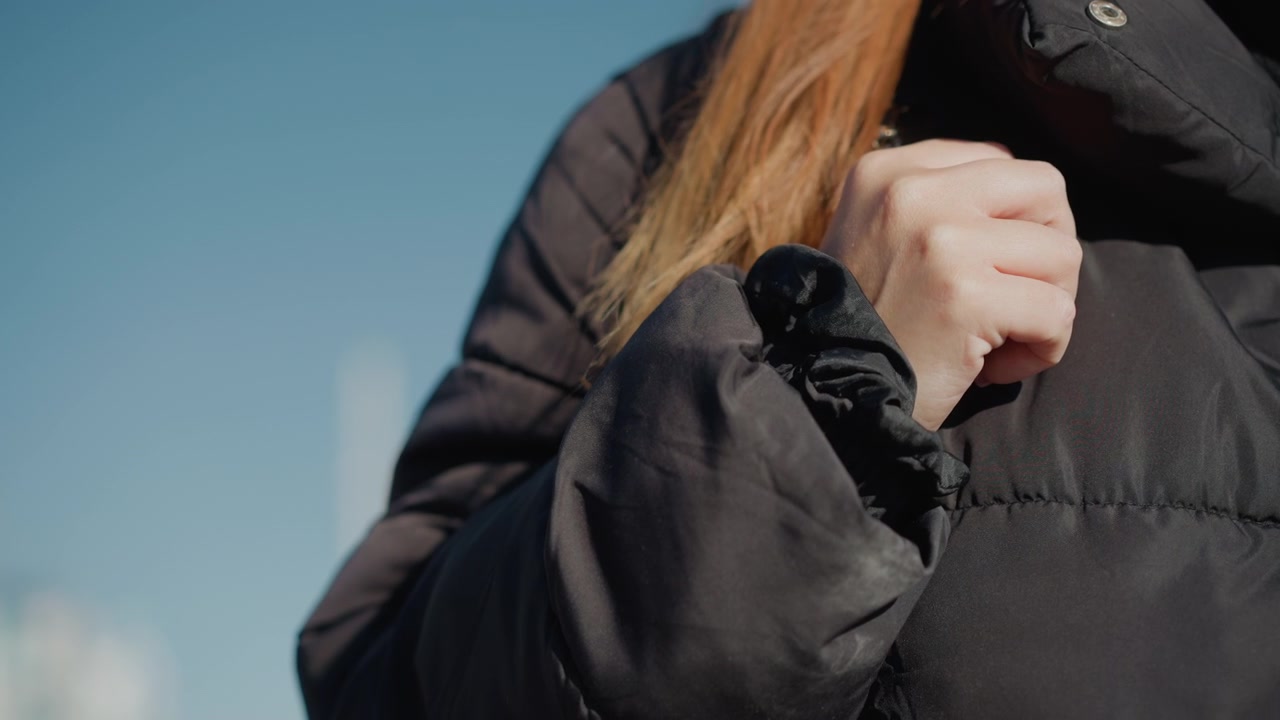 Donna regola la cinghia della sua giacca piumino nera contro uno sfondo di cielo azzurro limpido. I suoi capelli lunghi e la mano sono visibili mentre la donna sistema il capo con una luce naturale soffusa che evidenzia il tessuto.