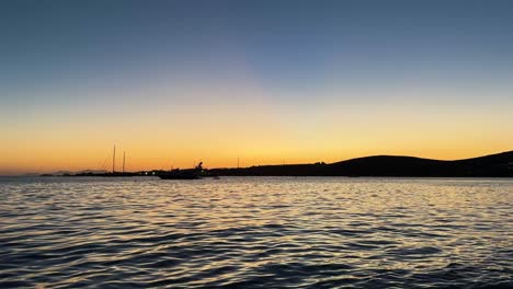 small boats and yachts on the cycladic sea in milos, greece, during a serene sunset with soft shadows, a blue sky, and dark silhouettes. static shot