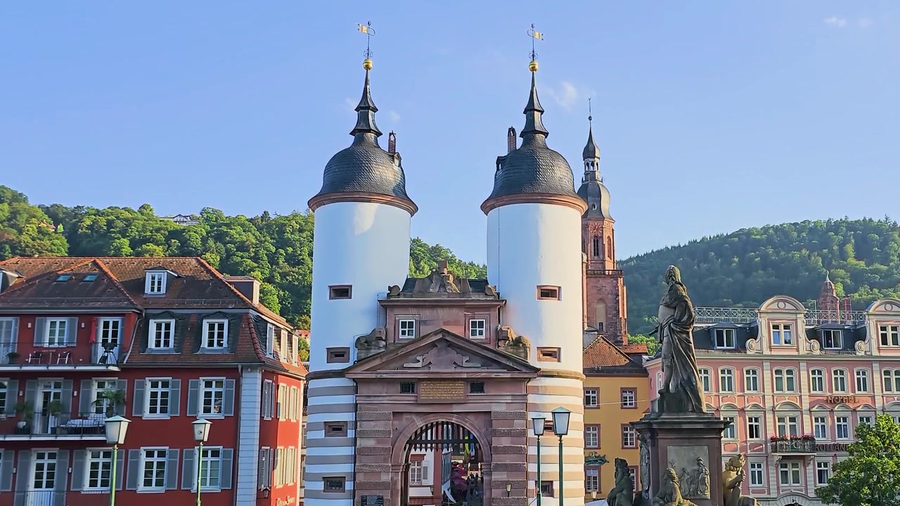 Heidelberg, Germania, vista del portale del Ponte Vecchio con due torri bianche gemelle, statua storica, edifici colorati del centro storico, guglia della chiesa e colline verdi sotto un cielo azzurro limpido nella luce dorata