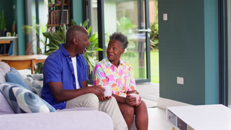 Smiling-Senior-Couple-Relaxing-On-Sofa-At-Home-Talking-And-Holding-Hot-Drinks