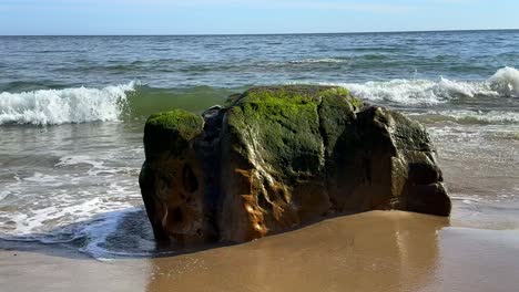 An-Einem-Sonnigen-Tag-Krachen-Wellen-An-Eine-Strandküste-Mit-Großen-Felsbrocken,-Die-Im-Sand-Stecken