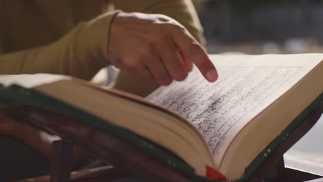 Primer-Plano-De-Una-Copia-Abierta-Del-Corán-En-El-Stand-En-Casa-Con-El-Hombre-Recitando-O-Leyendo-1