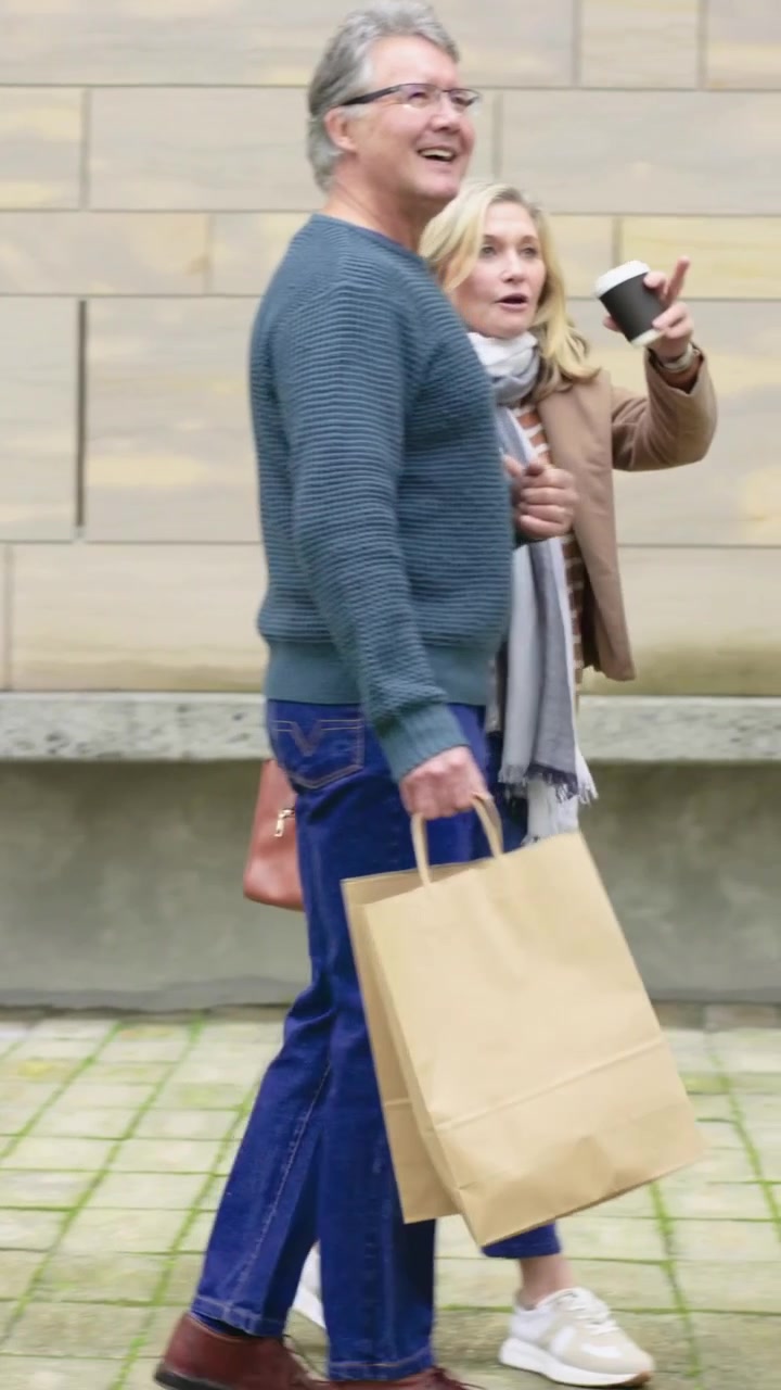 Video verticale: Coppia che entra in piazza, cammina a braccetto, porta una borsa, sorseggia caffè e fa shopping.