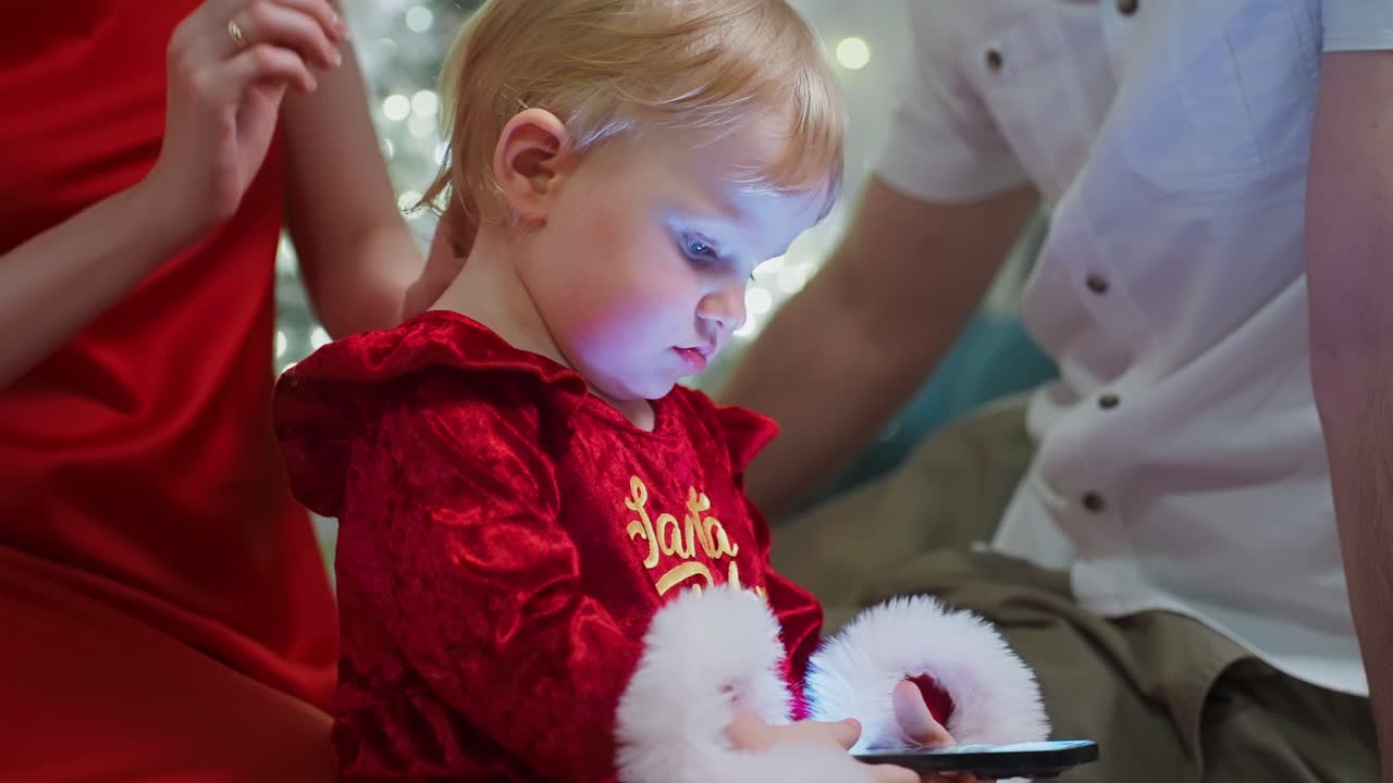 Child in festive attire focused on a smartphone, surrounded by family in a holiday setting, highlighting technology interaction during festive occasions