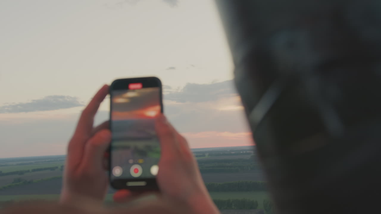 Close up rear view of lady capturing scenic sunset and flying paraglider on smartphone from hot air balloon, glowing sky and peaceful countryside visible in distance during golden evening light