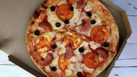 la pizza redonda fresca con tomates, salchichas, mozzarella y aceitunas en una caja de cartón gira lentamente.