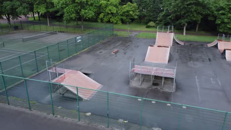 Vista-Aérea-Volando-Sobre-La-Rampa-Del-Parque-De-Monopatín-Cercado-Y-La-Cancha-De-Tenis-En-Un-Parque-Público-Vacío-Y-Cerrado