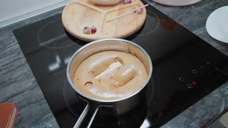 woman in plaid shirt drops butter into steaming pap as it melts and sinks, electric stove in modern kitchen with wooden tray and dry flowers in background