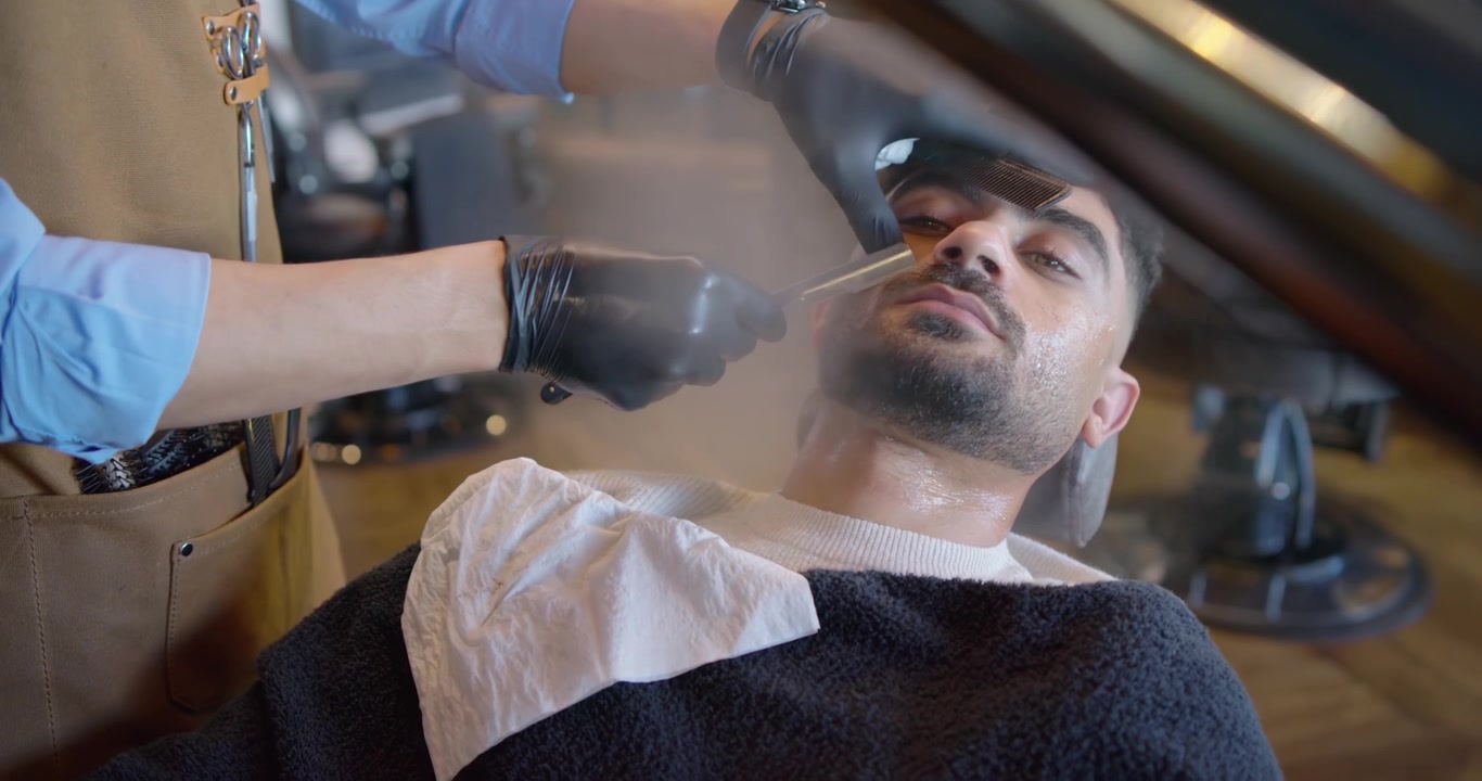 Mand læner sig tilbage i barberstol, frisør stabiliserer hovedet, påfører damp og trimmer skæg for at forme det.