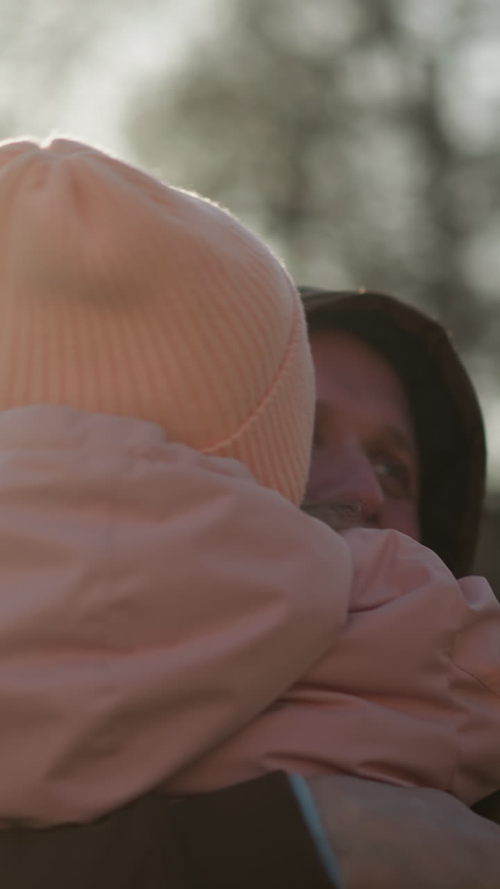 una niña con una gorra rosa y una chaqueta llevada por un hombre con una chaqueta marrón, mientras ella lo besa cariñosamente en la mejilla