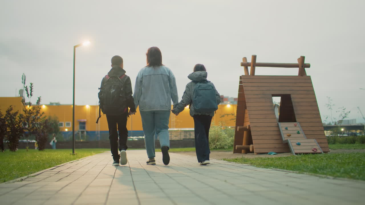 mum walking with two children holding hands down paved park path at dusk in urban green space, backpacks on shoulders, gentle evening stroll capturing family bond and support in relaxed outdoor setting