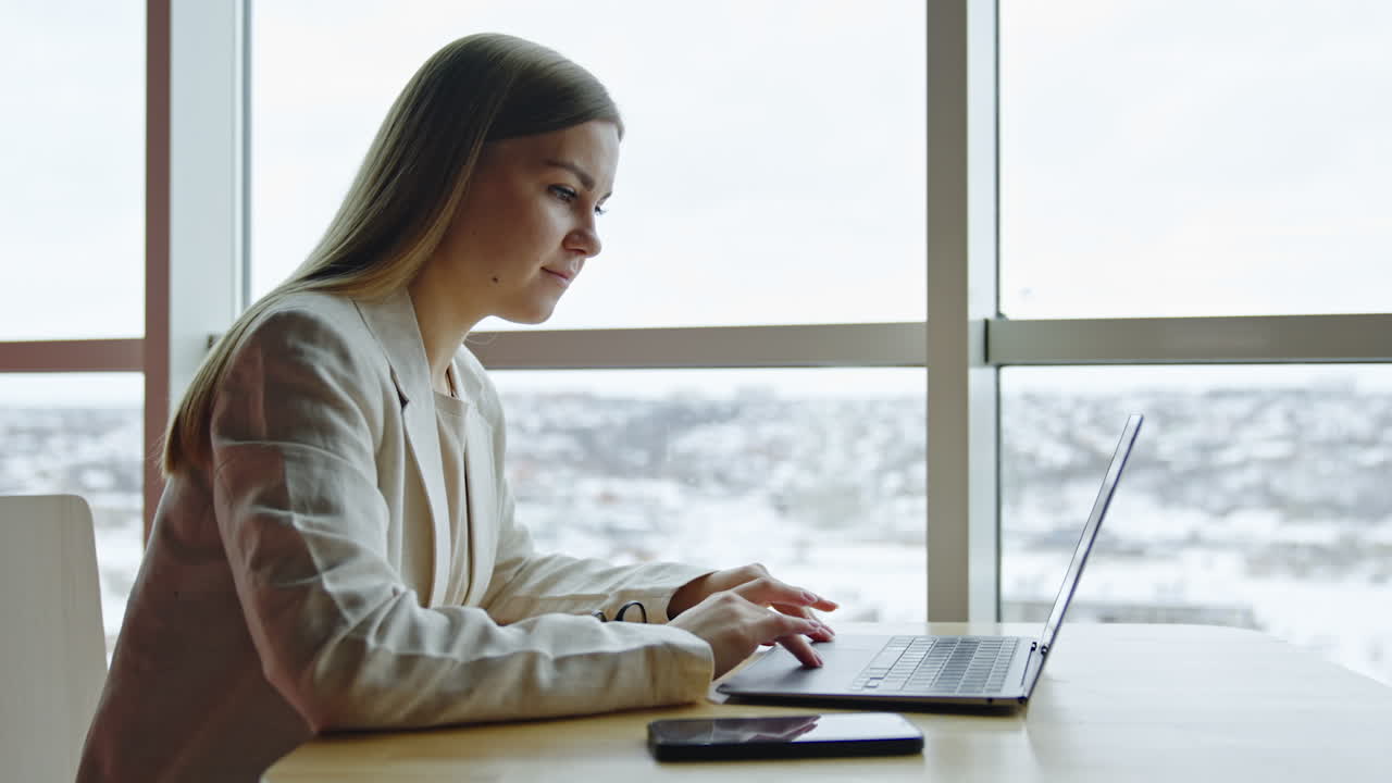 Long-haired woman working at her laptop. Blonde lady searches the web and smiles looking at the screen. Light windows at backdrop.