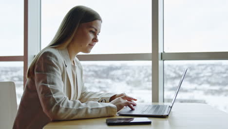 long-haired woman working at her laptop. blonde lady searches the web and smiles looking at the screen. light windows at backdrop.