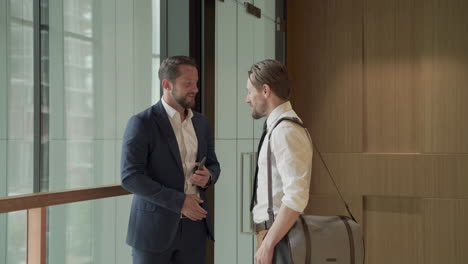 an attractive young businessman with a beard talks on the phone. a colleague arrives, greets him and they start talking