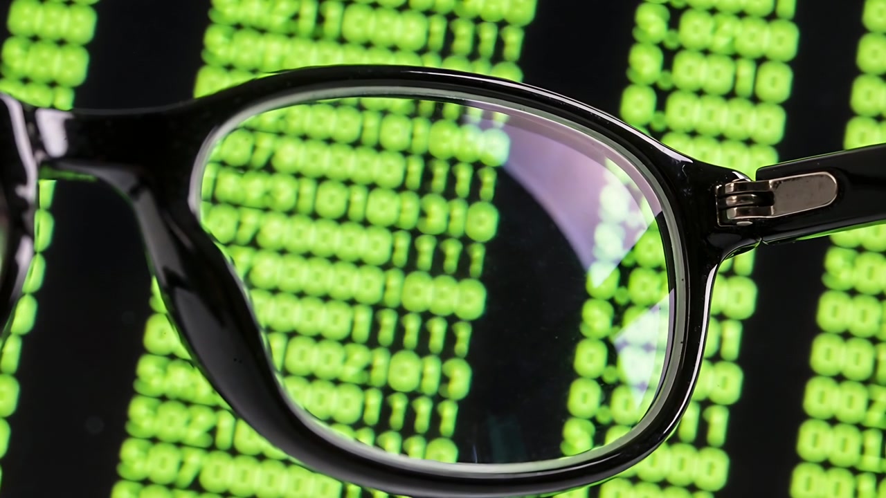 Showing black-rimmed eyeglasses magnifying scrolling green digits at workstation, with purple tint