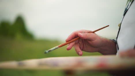 close-up shot of an artist s hand holding a paintbrush with a blurred palette visible in the background. the image captures the delicate grip and focus, set against a serene, blurred natural backdrop