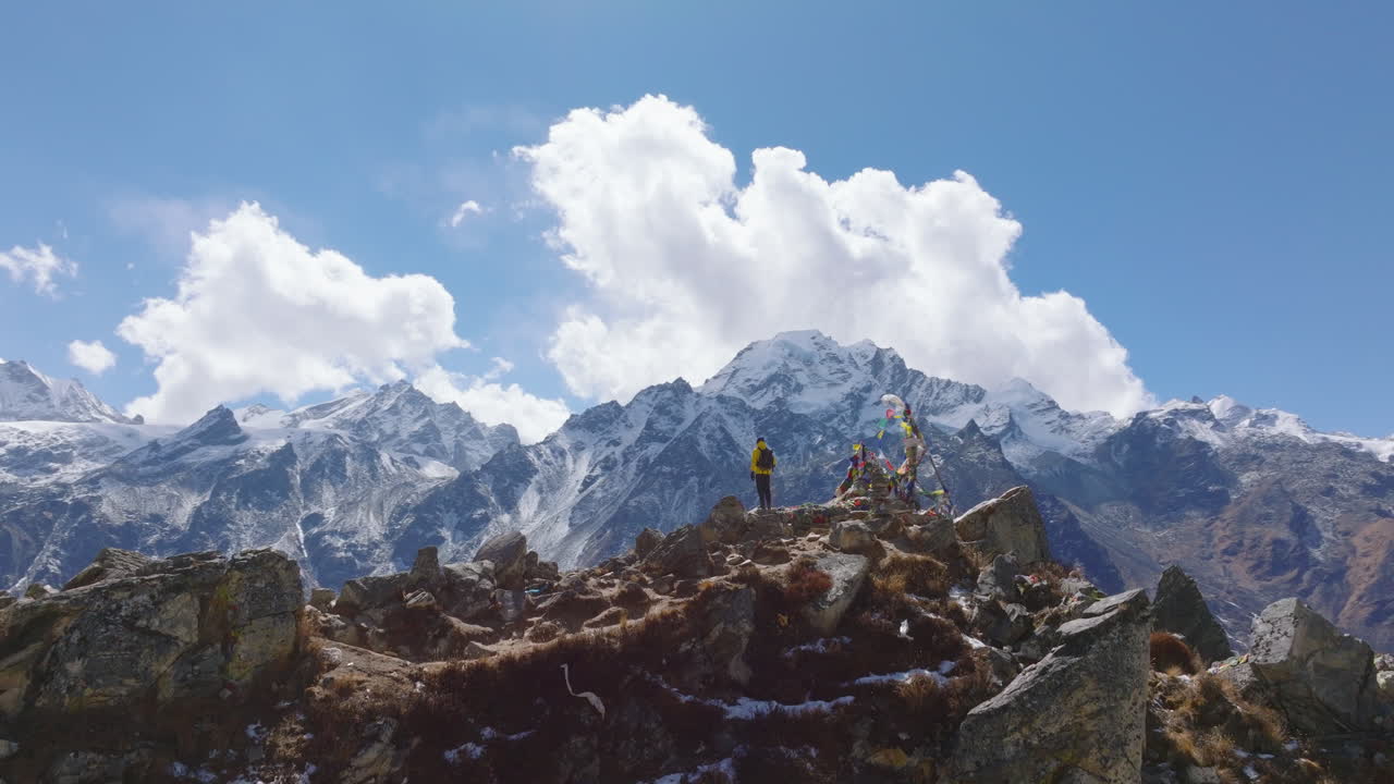 A male climber enjoys the summit view in Nepal with iconic mountains like Langtang and Everest. Captured by a drone, the scene features snow-capped peaks, vibrant prayer flags, and a clear blue sky.