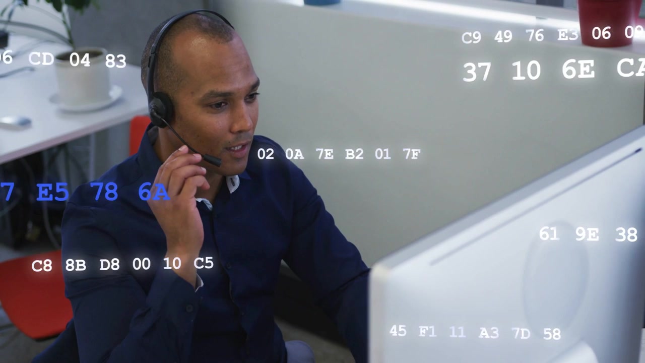 Mid-adult man receiving support call at desk, adjusting mic while hex codes scrolling on monitor