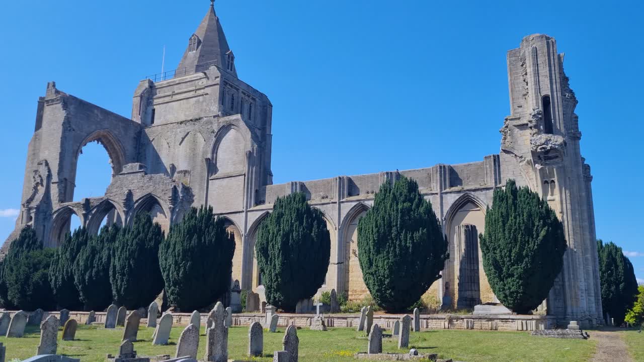 Revealing shot of the ruined side of Crowland Abbey in England, showing the historic monastery walls and cemetery in front under a clear blue sky on a sunny day