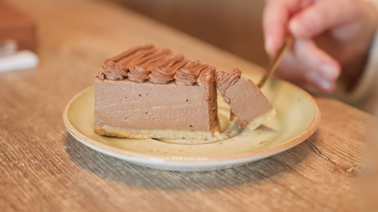 close up of person using fork to gently slice through creamy chocolate dessert on ceramic plate placed on wooden table surface, highlighting soft texture and inviting detail of sweet treat