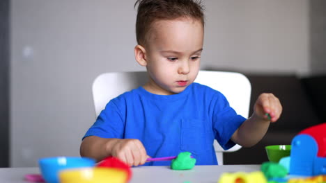 calm focused baby boy in blue t-shirt playing at desk. kid cuts the piece of green plasticine with a stick.