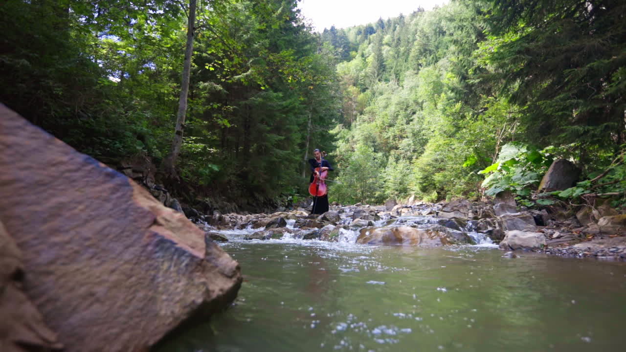 Beautiful woman with cello near the water. Female musician performing music at the river in the forest. Cellist plays the musical instrument in nature.