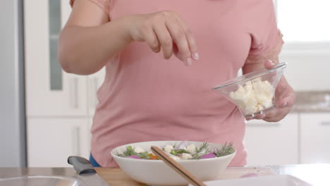 happy plus size biracial woman making feta cheese and vegetable salad in kitchen, slow motion