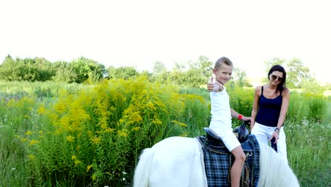 a woman and a boy are walking around the field, son is riding a pony, mother is holding a pony for a bridle. cheerful, happy family vacation. outdoors, in summer, near the forest