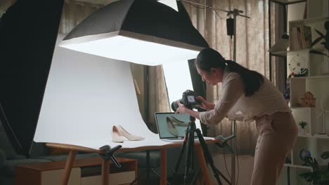 asian female photographer comparing photos on a laptop to camera while taking photos of women's shoes in home studio