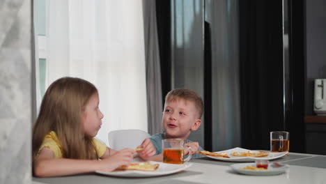 siblings enjoying breakfast together at dining table eating pizza slices drinking juice while little boy chats and laughs with sister filling cozy kitchen with happiness