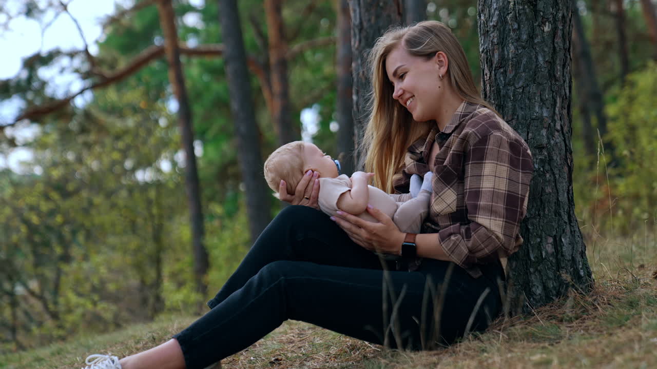 Happy first time mom speaking with her infant. Woman sits in the forest leaning on the tree and looking at her baby son.
