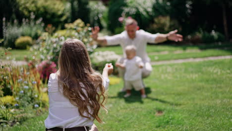 rear view of a long-haired woman calling up her little baby showing him a flower. kid stands near his dad and walks to flower bed.