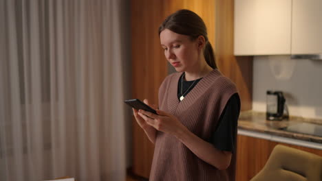 a young beautiful woman at home in the kitchen looks at her cell phone and drinks coffee. online messaging shopping and social networking