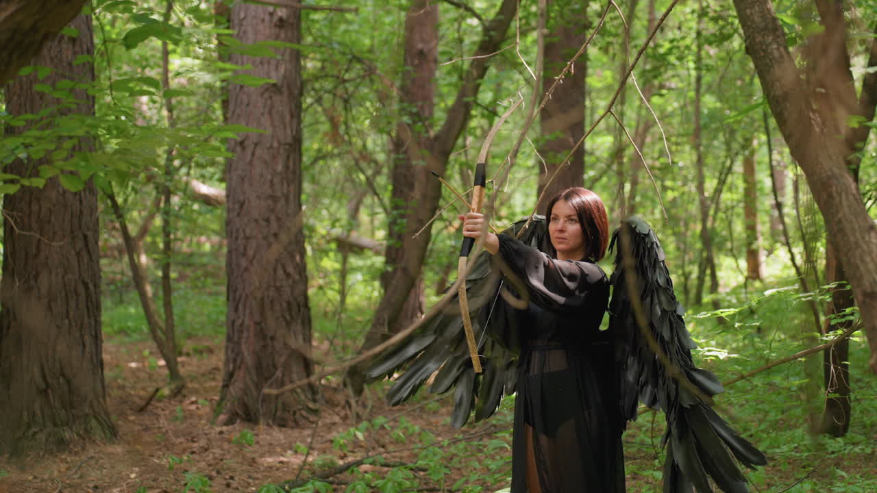 Woman dressed in black with large wings holding bow and arrow in forest, learning to shoot under soft sunlight, surrounded by trees and branches, atmosphere filled with focus