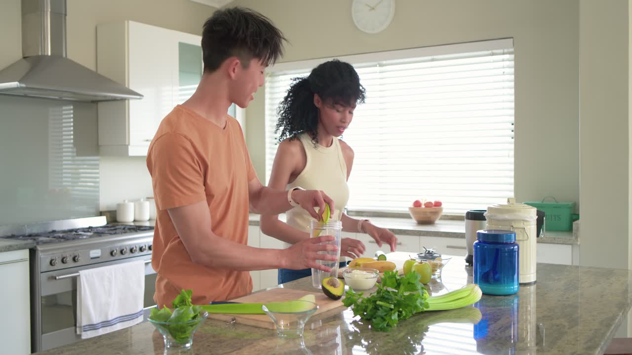 Casal diverso preparando abacate, cortando frutas em uma tábua e enchendo o liquidificador para fazer um smoothie