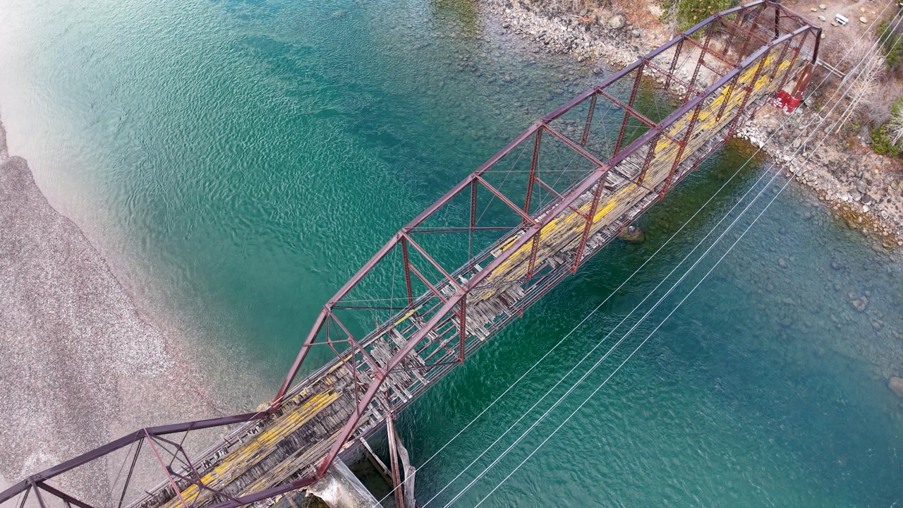 Aerial view of a suspension footbridge crossing the turquoise Flathead River near Whitefish, Montana, surrounded by rocky shores and forested landscape.