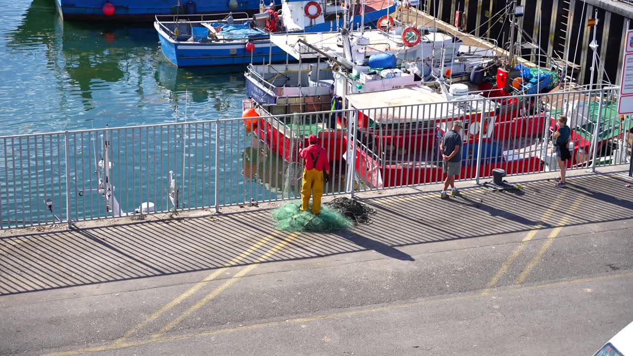 Worker in bright yellow overalls gathers fishing nets by docked boats at Santona port in Cantabria