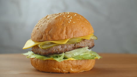 La-Hamburguesa-Gira-Sobre-Una-Tabla-De-Madera.-Una-Hamburguesa-Con-Ensalada-De-Chuleta,-Queso-Y-Tomates-Gira-Contra-Una-Pared-Gris