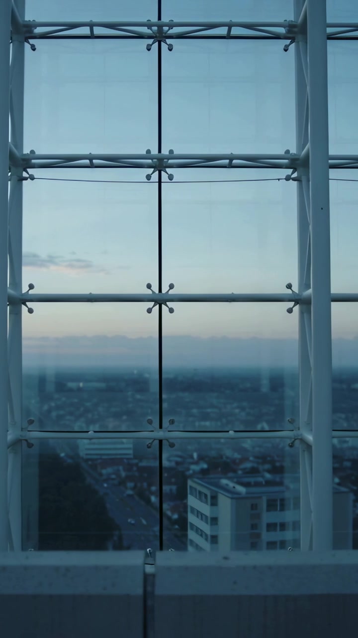 Video vertical: Cámara en movimiento que revela la parte baja de la ciudad a través de una ventana con parteluz al atardecer, dejando ver los tejados