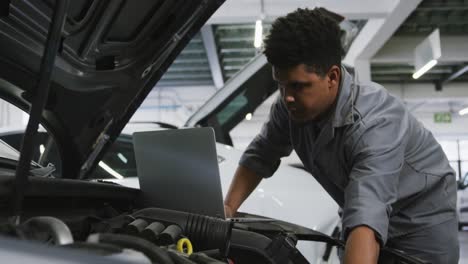 mecánico de automóviles afroamericano mirando un motor de automóvil abierto y usando una computadora portátil
