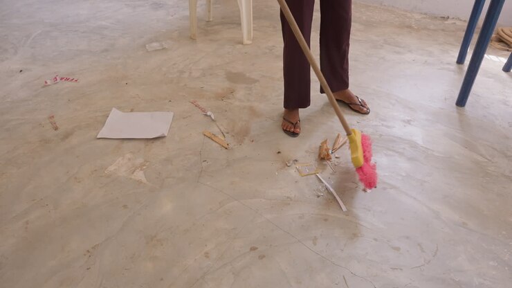Mocio che spazza briciole sparse su un pavimento di cemento, testa del mocio rosa e manico in legno, sedia di plastica e interni semplici, movimento di pulizia a piedi nudi, rapida sistemazione domestica dopo una fuoriuscita di snack, neutro.