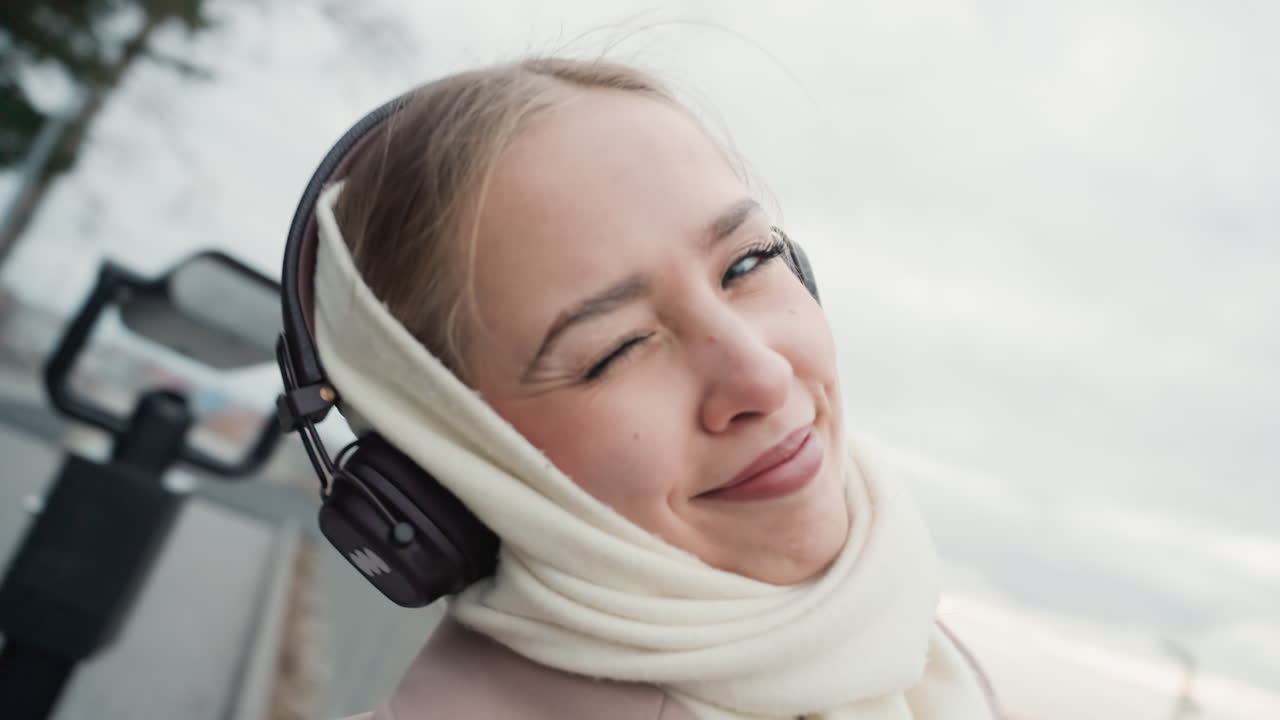 Ritratto urbano di donna gioiosa accanto al fiume. Donna con le cuffie che sorride vicino al lungomare in abbigliamento invernale. Immagine ravvicinata di donna allegra in cappotto e sciarpa che si gode la scena invernale urbana sull'acqua.