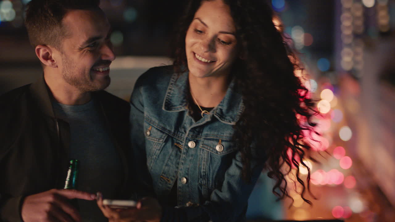 happy caucasian couple relaxing on rooftop at night browsing photos using smartphone enjoying romantic date in city at night