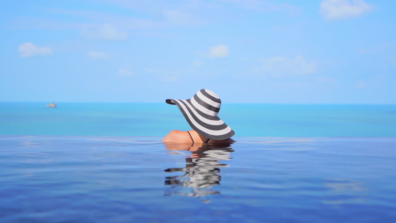 Back View of the Young Woman in Infinity Swimming Pool wearing a Large Black and white Hat and leaning her Arms on the Edge of the Pool in Spain